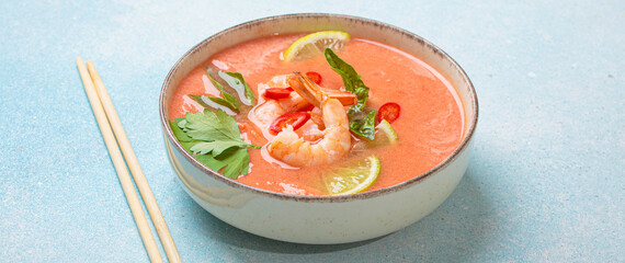 Creamy flavourful tom yum soup, a popular dish of Thailand, is served in a bowl, garnished with fresh shrimp, lime wedges, herbs, and chili peppers, accompanied by chopsticks on a linen napkin