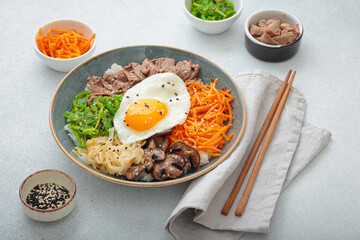 Bibimbap, authentic korean dish with a steamed rice topped with fried egg, beef, colorful vegetables, traditional dish of Korea on light background with chopsticks
