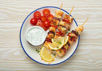Grilled chicken skewers seasoned with herbs and spices, served on a plate with lemon wedges, cherry tomatoes and yogurt sauce, flavorful and healthy meal mediterranean style on white wooden background