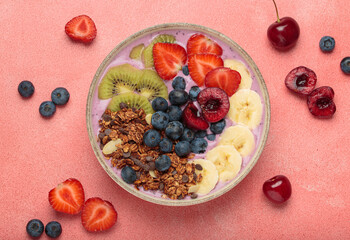Vibrant and nutritious acai bowl topped with sliced kiwi, strawberries, blueberries, cherries, banana, and granola, a colorful and healthy meal or breakfast on a pink background