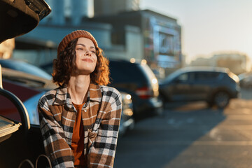 Fototapeta premium Woman in beanie and plaid jacket sits by open car trunk in parking lot, sunlight on her face, candid smile and golden hour glow conveying authenticity, mindful living and emotional storytelling