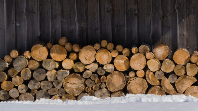 Stacked logs provide warmth and beauty against a rustic wooden backdrop in a winter landscape.