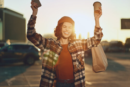 Woman holding coffee and paperbag smiling in urban street at golden hour glow, candid joyful shopping moment with phone raised, authenticity and mindful living in emotional storytelling scene.