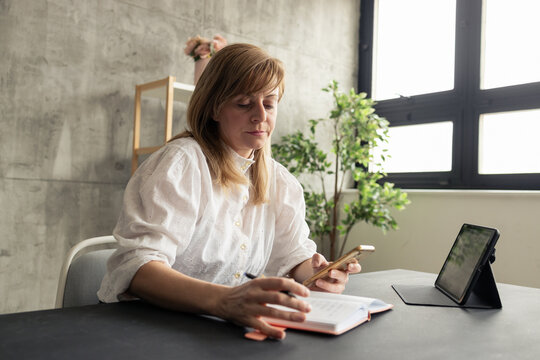 Businesswoman working from home using smartphone and taking notes on notebook - Powered by Adobe
