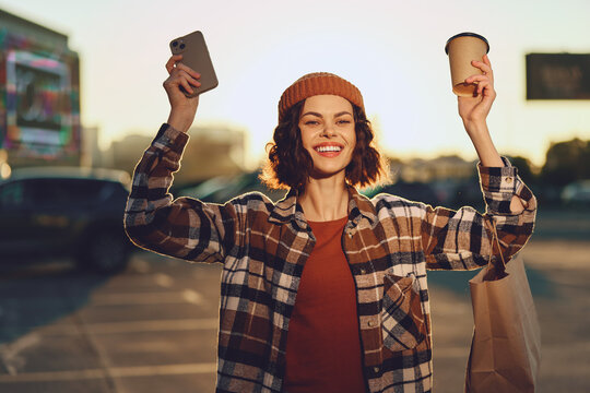 Woman holding coffee and phone with shopping bag, smile in urban golden light; authenticity and candid lifestyle capture golden hour glow, mindful living and emotional storytelling.