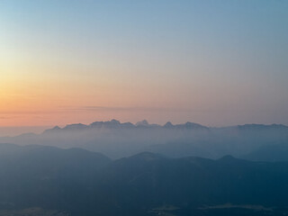 Layers of misty mountain silhouettes of the Julian Alps recede into the distance under a soft, gradient sky during a tranquil and atmospheric sunrise, as seen from Dobratsch in Carinthia, Austria.