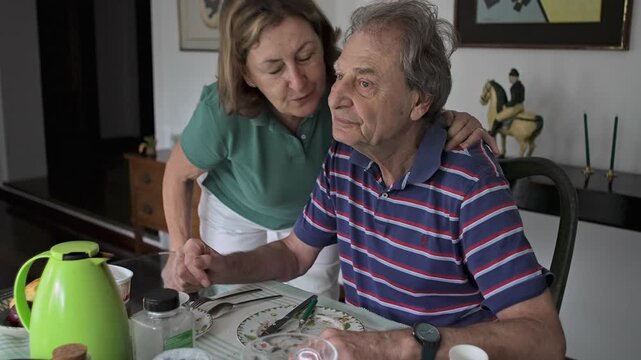 Elderly Woman Kissing Husband Goodbye As He Sits At Breakfast Table Before Leaving Home For Daily Routine