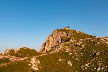 A winding hiking trail meanders through a grassy alpine meadow, leading the eye towards the iconic Maria am Stein church which sits majestically on rocky summit against clear blue sky at first light.