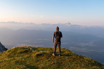 A lone hiker with a backpack stands on the summit of Dobratsch, watching the beautiful morning sunrise over the majestic Julian Alps and the city of Villach in Carinthia, Austria, enjoying the view.