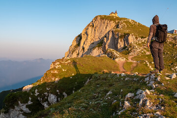 A lone hiker with a backpack stands on a rocky outcrop, gazing at the Maria am Stein chapel perched on sunlit peak, ready for the final ascent on Villacher Alpe against vast mountain backdrop at dawn.