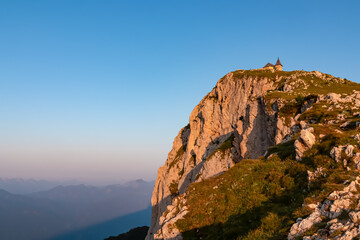 The historic Maria am Stein chapel sits atop a dramatic, sun-kissed rock cliff on the Villacher Alpe, its golden walls glowing in early morning light against a clear blue sky and a hazy alpine vista.