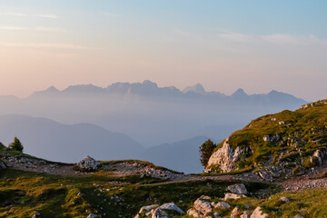 A rugged alpine hiking trail winds through a grassy, rocky meadow on the Villacher Alpe, leading towards breathtaking panoramic view of distant, mist-shrouded Julian Alps under a soft pastel dawn sky.