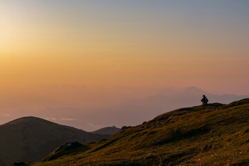 Lone hiker meditates on a grassy slope of the Villacher Alpe at dawn, their silhouette gazing at vast, serene panorama of hazy mountains and glowing river valley under golden sunrise sky in Carinthia.