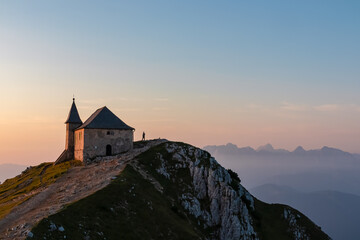 Solitary figure stands near the ancient Maria am Stein chapel on the summit of the Villacher Alp (Dobratsch), taking in the epic panoramic view as the sun rises over the misty Julian Alps in Austria.
