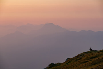 Lone hiker sits in quiet contemplation on a grassy mountain slope on Dobratsch, their silhouette dwarfed by vast and serene vista of hazy, layered alpine ranges under a soft pastel-colored sky at dawn