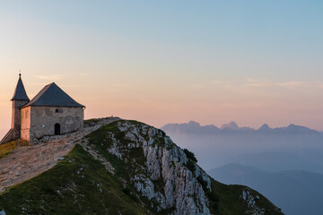Historic Maria am Stein chapel is warmed by the first light of dawn on the Dobratsch summit, its ancient stone walls set against a majestic and misty panorama of the Julian Alps in Carinthia, Austria.