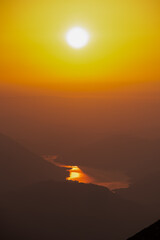 Brilliant morning sun hangs high in a golden sky, its intense light casting shimmering, molten gold reflection upon winding river nestled deep within hazy, silhouetted valley as viewed from Dobratsch
