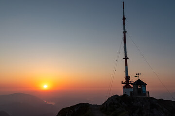 Dobratsch transmission tower and a weather station are silhouetted against a brilliant sunrise in Austria, overlooking hazy valley with a distant river, blending technology with natural alpine beauty