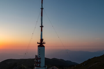 Tall red and white telecommunications tower stands prominently on the Dobratsch summit, captured from high angle against a breathtaking sunrise with clear sky grading from orange to blue over Austria