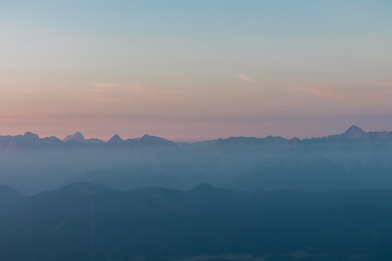 The distant, jagged peaks of the Julian Alps create a stunning panoramic silhouette, emerging through layers of dense blue fog and morning mist under a soft, beautifully colored pastel sky at sunrise.