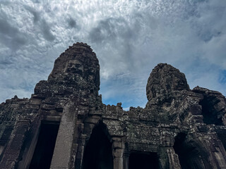 Two giant, weathered stone towers featuring serene carved faces loom majestically against dramatic and moody cloudy sky, capturing powerful atmosphere of Bayon temple in Angkor, Siem Reap, Cambodia