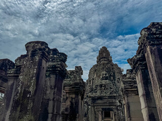 Weathered stone pillars stand before a magnificent tower with a carved serene face, reaching towards a dramatic cloudy sky at the historic Bayon temple in the Angkor Thom complex, Siem Reap, Cambodia.