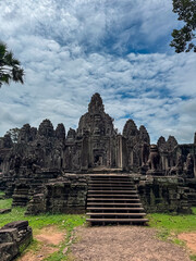 Central towers of the ancient Bayon temple rise under a vast, cloudy sky, with stone lion statues guarding the entrance stairs at the historic Angkor Thom archaeological park in Siem Reap, Cambodia.