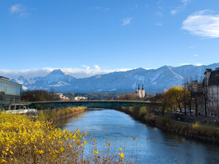 Fototapeta premium Villach, Austria - November 18, 2025: Scenic view of the river with autumn foliage, mountains in the background, and a historic church, capturing the beauty of nature and architecture