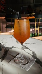 A refreshing orange cocktail, likely an Aperol Spritz, in a wine glass with a straw and orange peel garnish, resting on a white marble table in a stylish bar setting