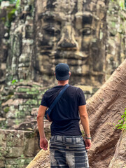 A male traveler, seen from behind, contemplates the enigmatic, out-of-focus stone face of a historic temple gate, creating a mysterious and personal travel moment in Angkor, Siem Reap, Cambodia.