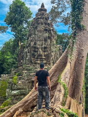 A young male tourist stands in awe before a giant ancient stone face tower at a gate of Angkor Thom, framed by massive tree roots, exploring the historic temple complex in Siem Reap, Cambodia.
