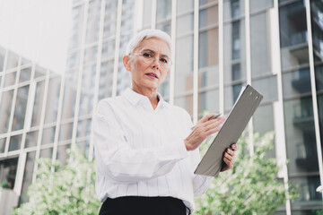 Confident elderly businesswoman stands outdoors in front of glass city buildings using a tablet for a modern professional image