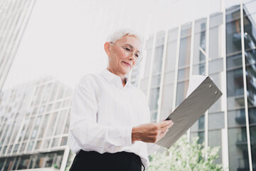 Senior businesswoman stands outdoors in front of modern city buildings holding a clipboard and smiling confidently