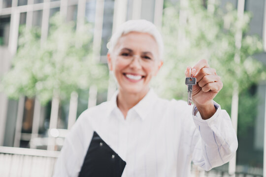 Confident elderly woman businesswoman outdoors smiling and presenting keys in a modern city setting as she stands in daylight wearing a white shirt and holding a key ring - Powered by Adobe