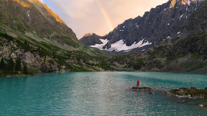 a man stands on an island, a drone flies over the turquoise-colored mountain lake of Kuyguk, and there is a rainbow in the sky