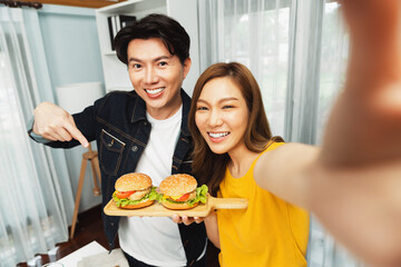 Smiling host chef influencers taking photo with smartphone by selfie straight view holding double hamburger on cooking show special easy dish homemade on social media channel live streaming. Infobahn.