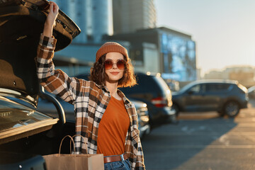 woman shopping by car trunk at sunset in urban parking, portrait of beanie style with sunglasses...
