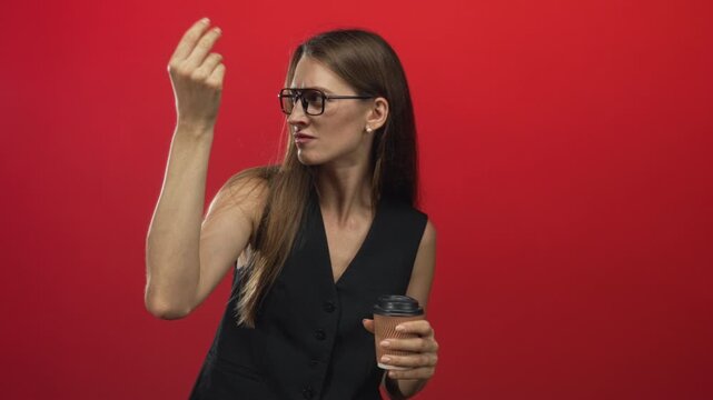 Woman wearing glasses and black vest holding a paper coffee cup and gesturing with hand in red studio; annoyance.