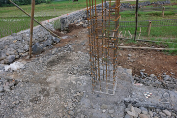 A close-up view of a reinforced steel column frame partially covered in fresh concrete at a house construction site, showing the early stage of foundation and structural work.