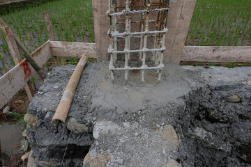 A close-up view of a reinforced steel column frame partially covered in fresh concrete at a house construction site, showing the early stage of foundation and structural work.