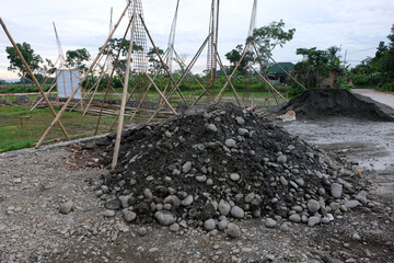 A pile of construction materials, including gravel and soil, at a house-building site with reinforced steel columns and wooden supports visible in the background.
