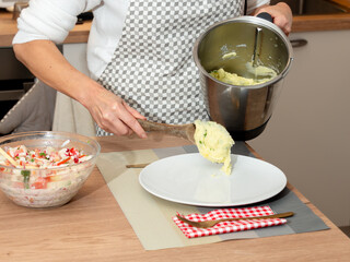 A woman serves freshly prepared mashed potatoes onto a plate. A bowl of vegetable salad is visible in the background. Home cooking and meal preparation.
