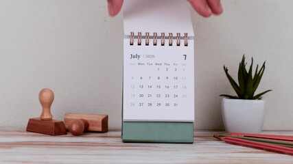 Hand Adjusting Desk Calendar Displaying July 2026 Beside Office Supplies