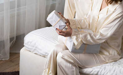 Woman in cream satin pajamas on cozy bed holding neatly wrapped gift boxes. Calm, intimate holiday atmosphere. Greetings and celebration. Joy of giving and receiving presents at home.