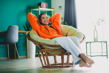 Relaxed woman in bright orange sweater sits in a wicker chair at home enjoying leisure and comfort...