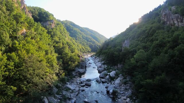 Backlit aerial flight moving forward above the rocky riverbed of the Arda River, revealing large boulders, steep slopes, and dense green trees on both sides