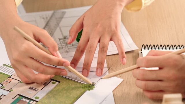 Architects using measurement tool and pencil tracing in building blueprint by coworker support on table with architectural document and wooden block scatter around at office. Closeup. Delineation.