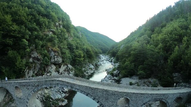 Drone flying backward over the Arda River gorge, rising toward the stone arches and smoothly passing above the Devil&rsquo;s Bridge at sunset