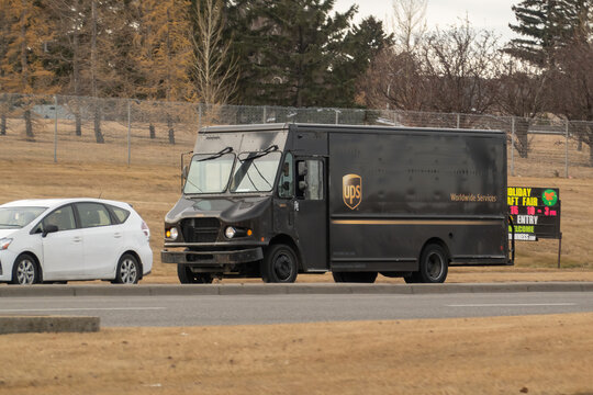 Calgary, Alberta, Canada. Nov 18, 2025. A brown UPS package delivery truck drives on an airport service road with a parked, branded UPS cargo jet visible in the distance.