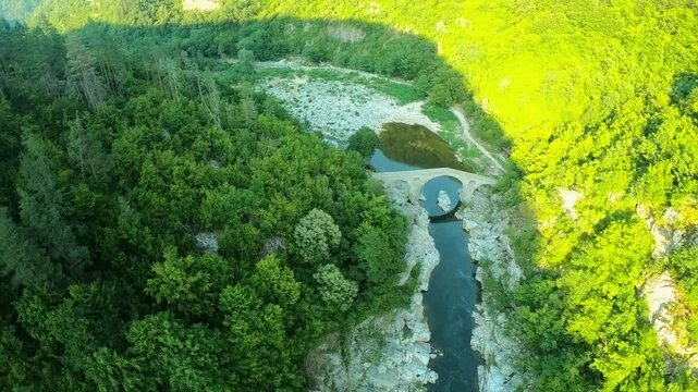 High-altitude sunset drone shot over the Devil&rsquo;s Bridge and the Arda River. The camera moves backward, gradually pulling away from the historic stone bridge and revealing the dramatic Rhodope mountain